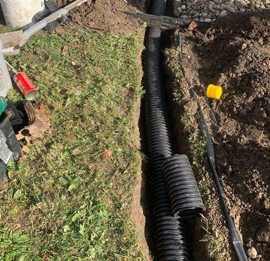 A narrow trench with black corrugated drain pipe installed. The trench is in a grassy area with dirt.