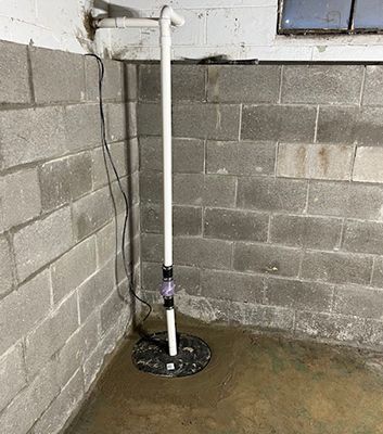 A sump pump in a basement. The pump is black with a white pipe leading up to the ceiling. Concrete block walls.