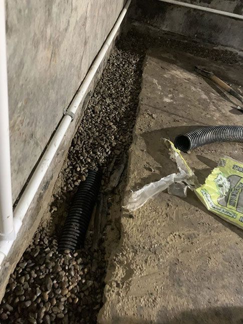 Basement corner with gravel-filled trench. Black drain pipe, white pipe, and torn packaging visible.