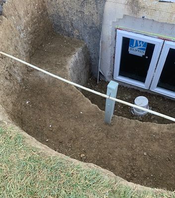 Excavated area around basement window; dirt walls and floor. Pipe and water tank near window; grass at edge.