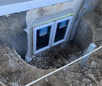 A window installed in a foundation wall, surrounded by dirt and construction materials.