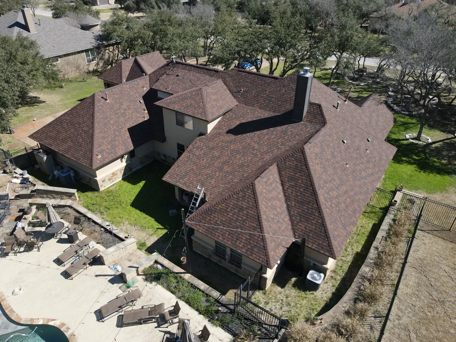 Aerial view of a brown-roofed house with a swimming pool and yard, surrounded by trees.