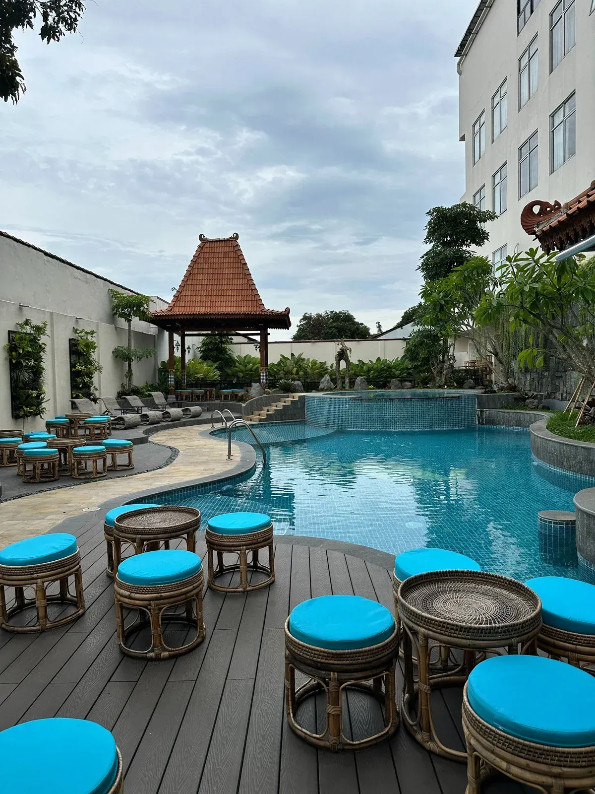 Pool area with blue stools, gazebo, and a waterfall. Hotel building in the background, overcast sky.
