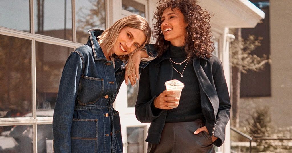 Two women lean together outside. One in denim jacket, the other with drink.