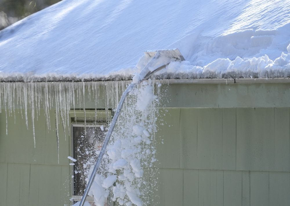 A roof rake pulls a large chunk of snow off a snow-covered roof, with icicles hanging from the gutter above a window.