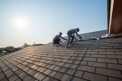 Two workers wearing safety helmets install gray roof shingles on a residential building under a bright, sunny sky.