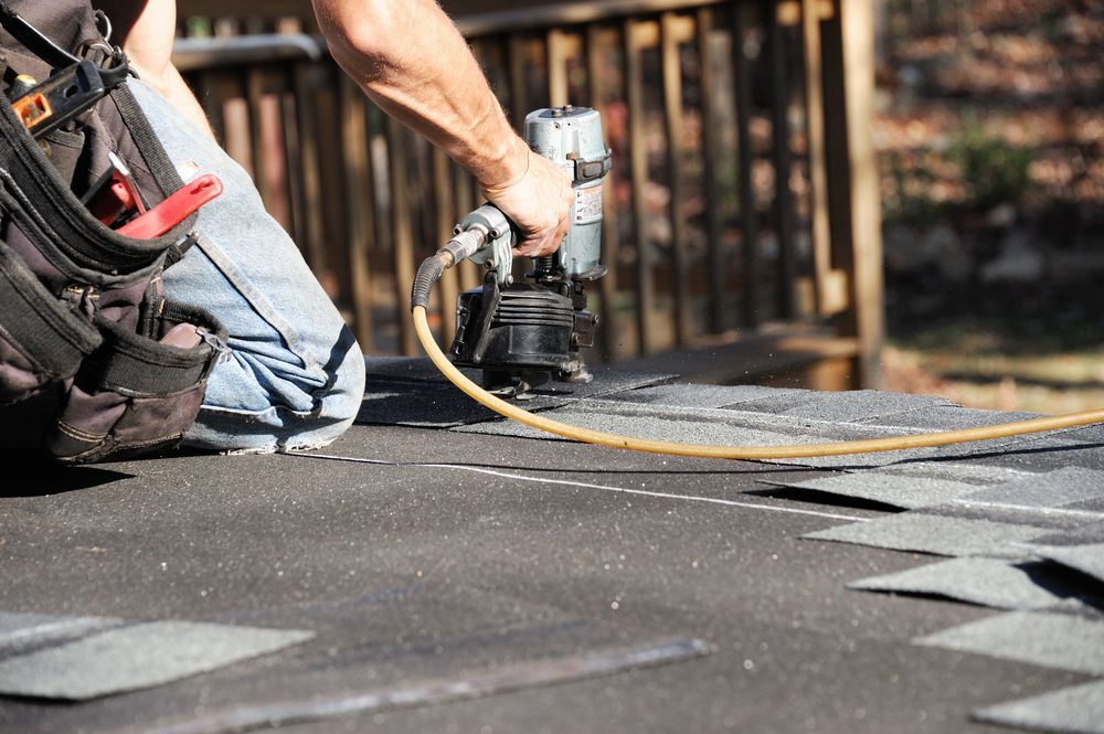A person kneels on a roof, using a pneumatic nail gun to install dark asphalt shingles.