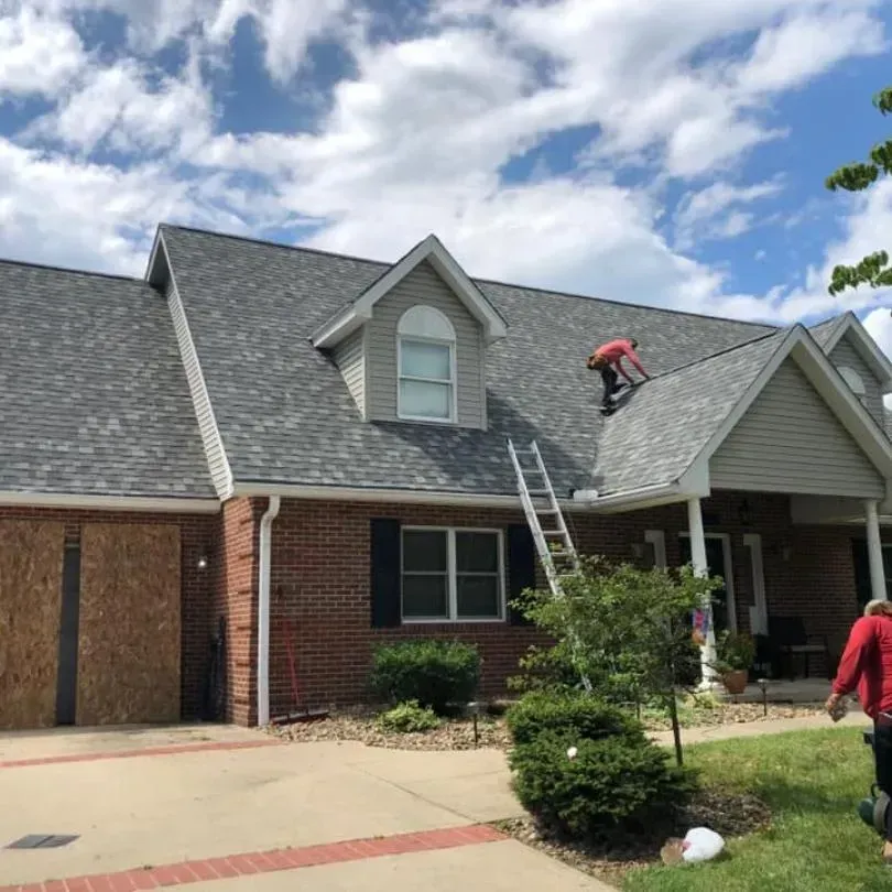 A person in a red shirt works on the grey shingled roof of a brick house with a ladder leaning against it.