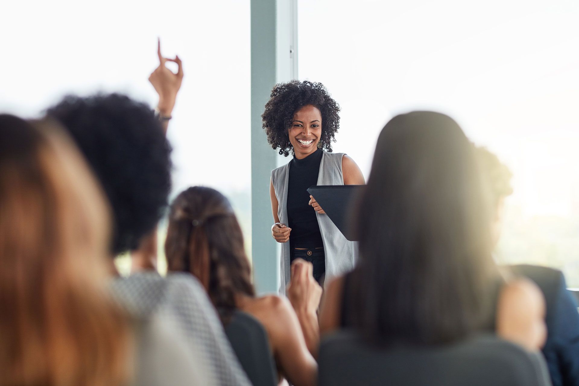 Woman presenting to an audience, smiling. Someone in the audience raises a hand.
