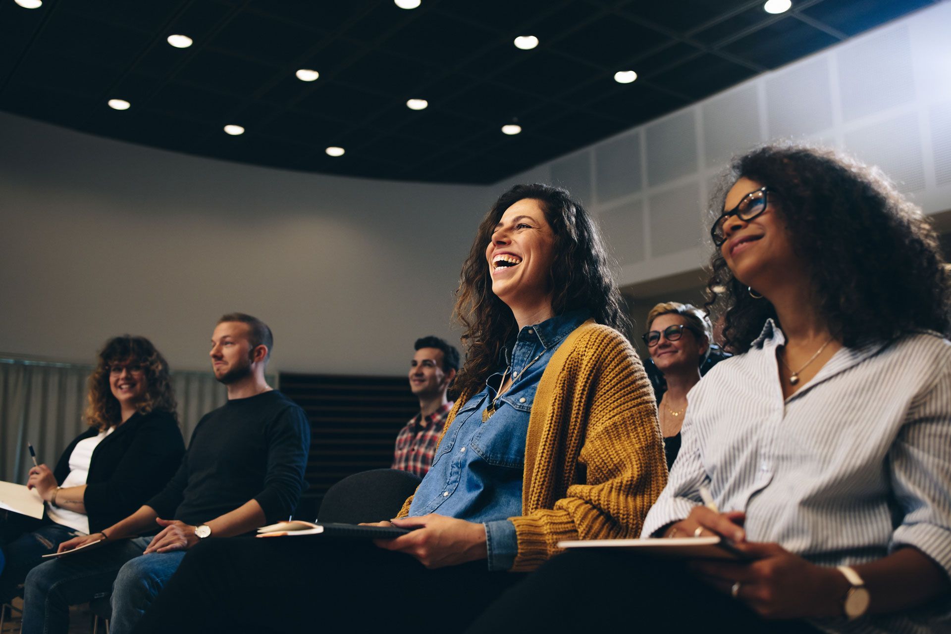 Audience laughs during a presentation in an auditorium; attendees hold notebooks, some smiling.