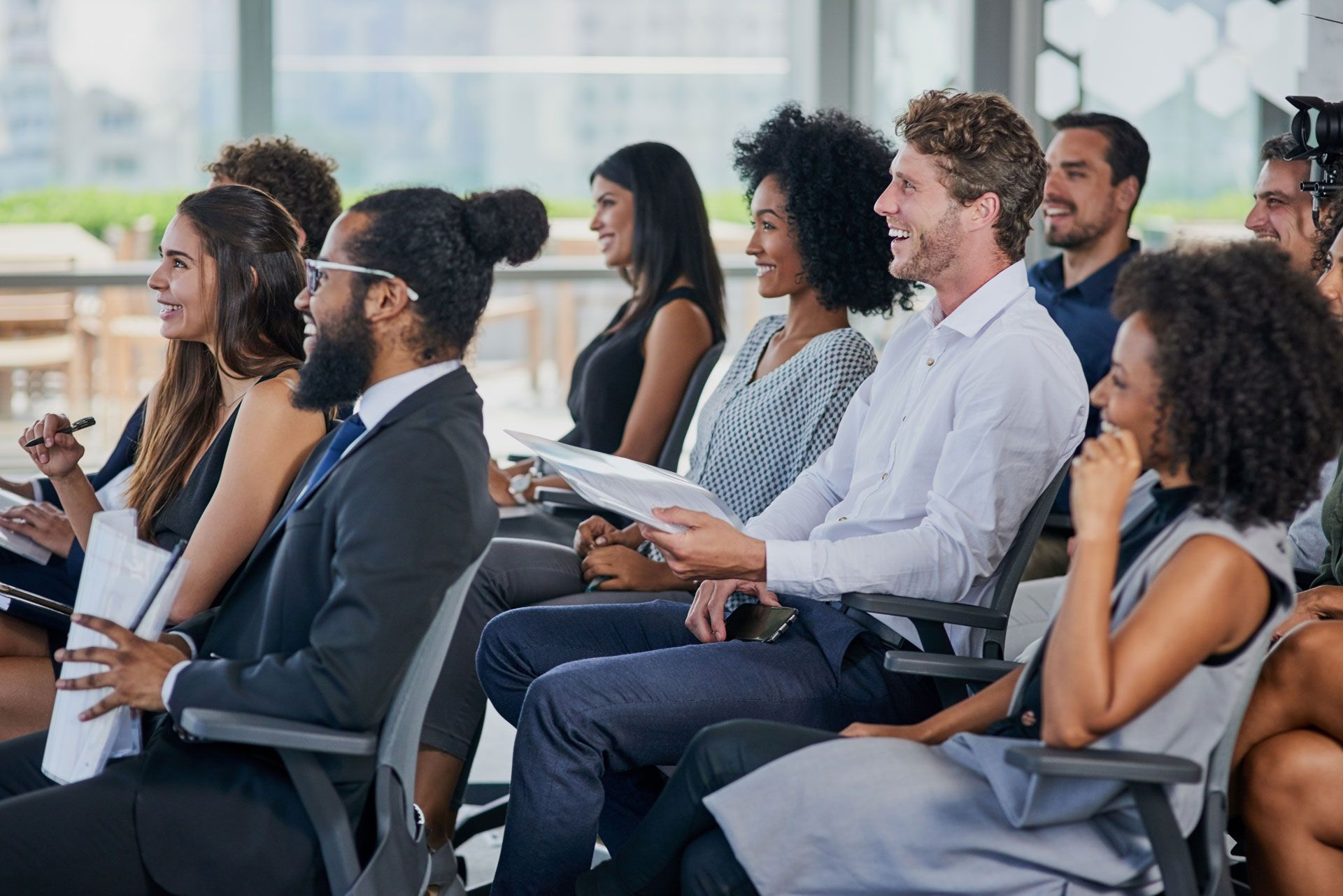 People in business attire seated, listening and smiling at a presentation in a modern setting.