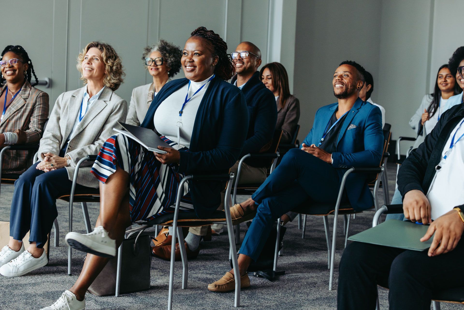 Audience members in a conference room smiling. People seated and holding folders, attending a presentation.