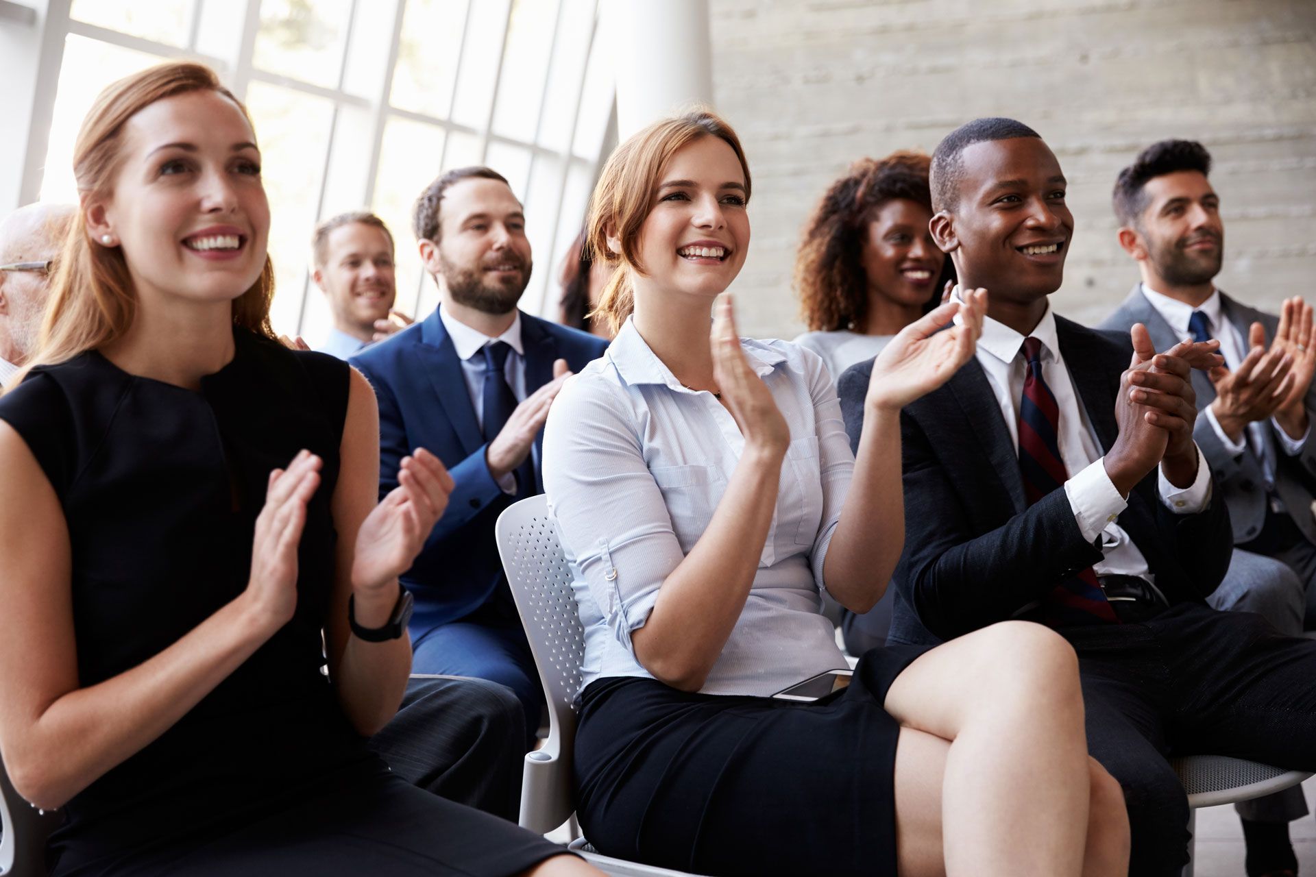 People in business attire clapping in an audience, smiling. Inside a modern building.