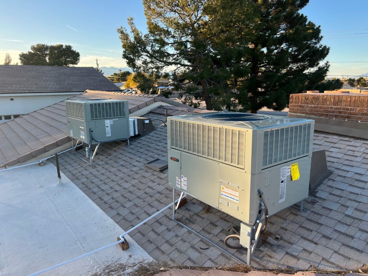 Two air conditioning units on a rooftop. The units are gray, mounted on metal stands, with a brown shingle roof.