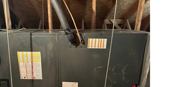 HVAC unit in an attic. Gray metal box with vents and labels. Black vent pipe and wires overhead.