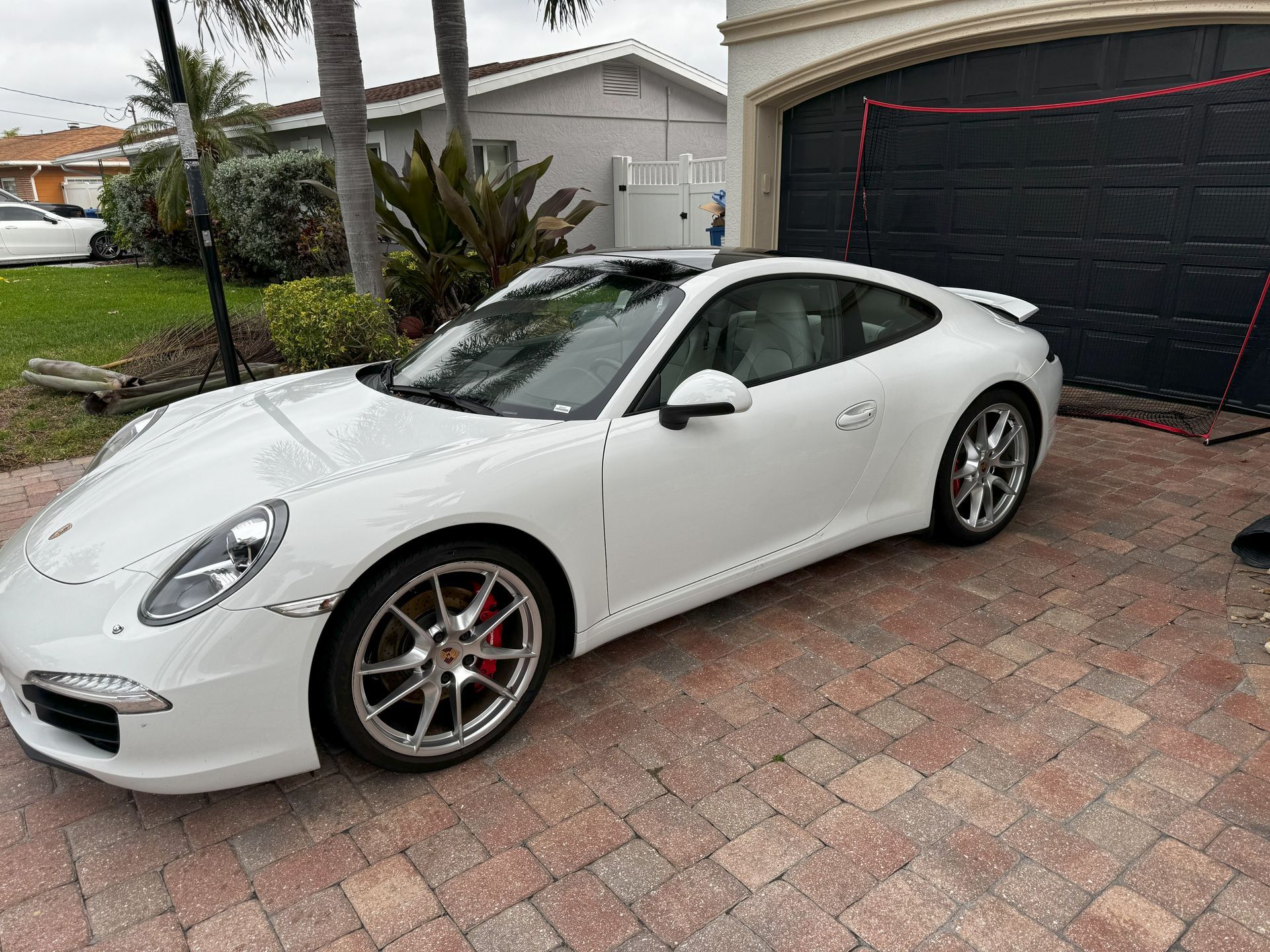 White Porsche sports car parked on a brick driveway in front of a garage and house.