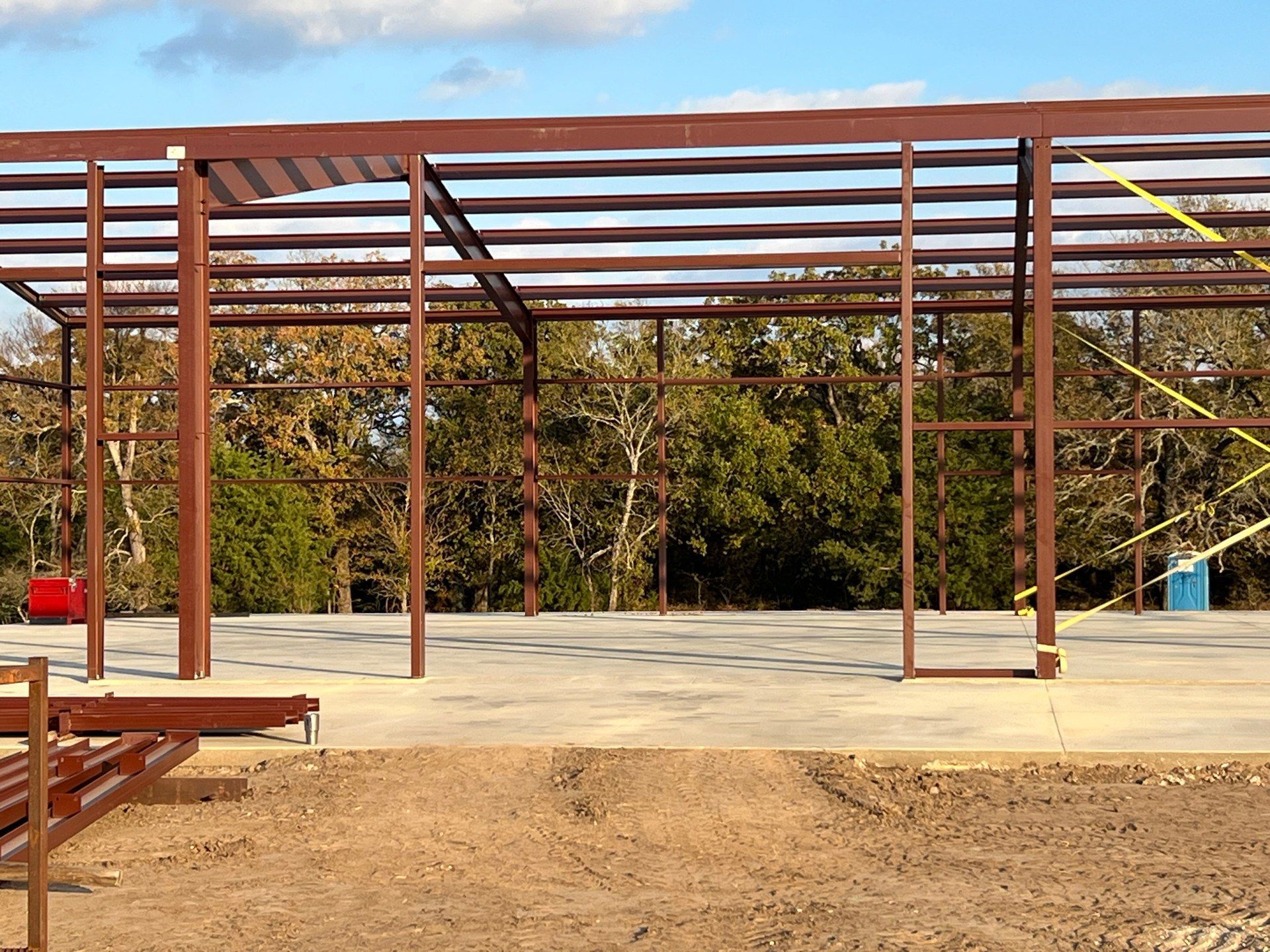 The structural steel frame of a building under construction on a concrete foundation, set against a background of trees.