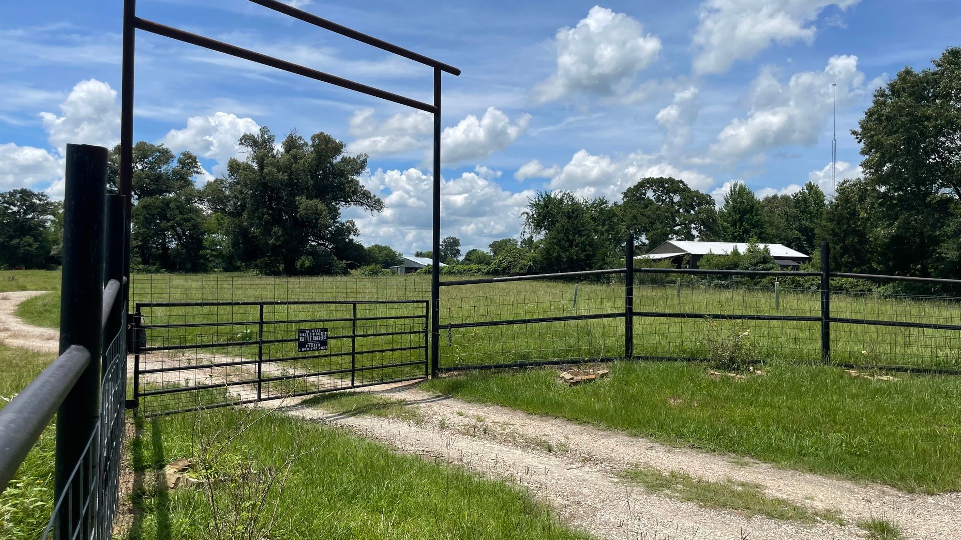 A metal gate entrance leads to a grassy field with a dirt path, surrounded by trees under a partly cloudy blue sky.