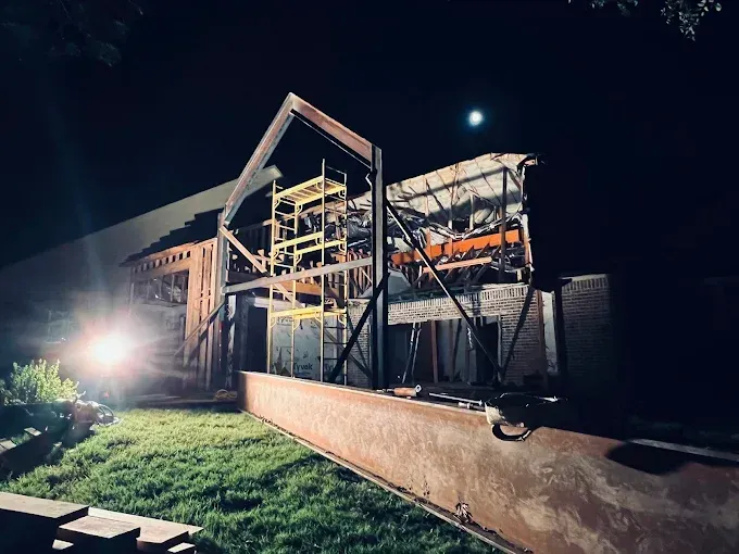 A steel frame for a building extension stands illuminated at night, with yellow scaffolding beside a grass yard.