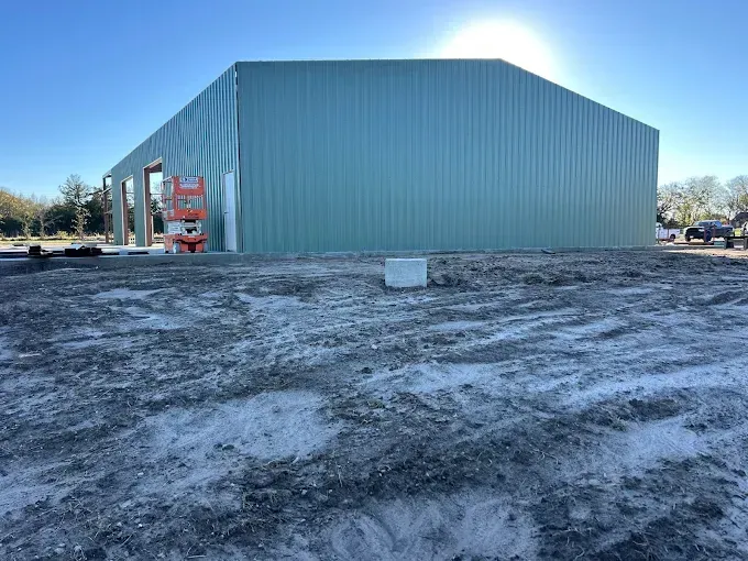 A light green metal building under construction on a dirt lot with a construction lift visible by the front entrance.