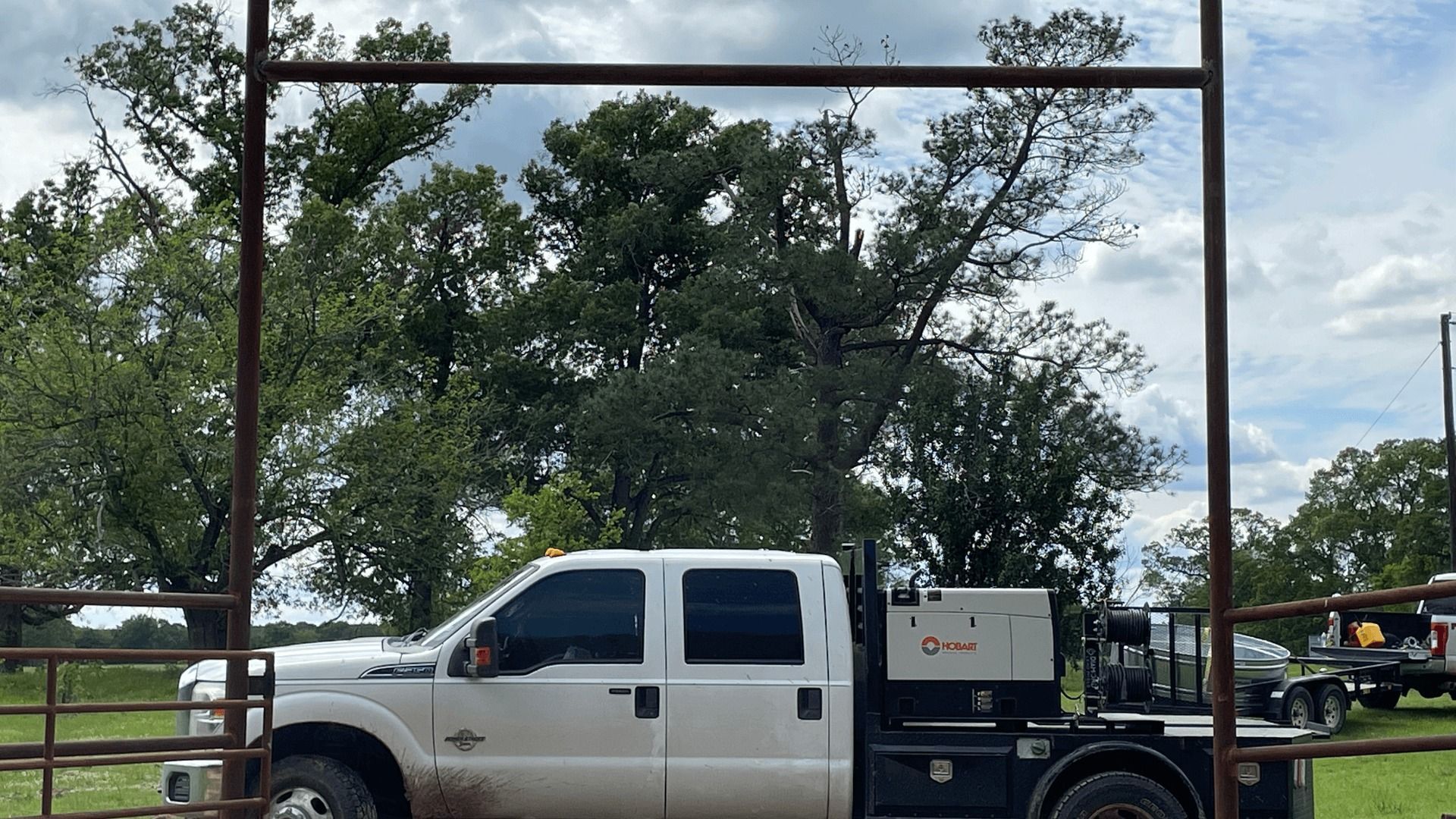 A white utility truck with a service bed is parked outdoors, framed by a rusted metal gate under a cloudy sky.