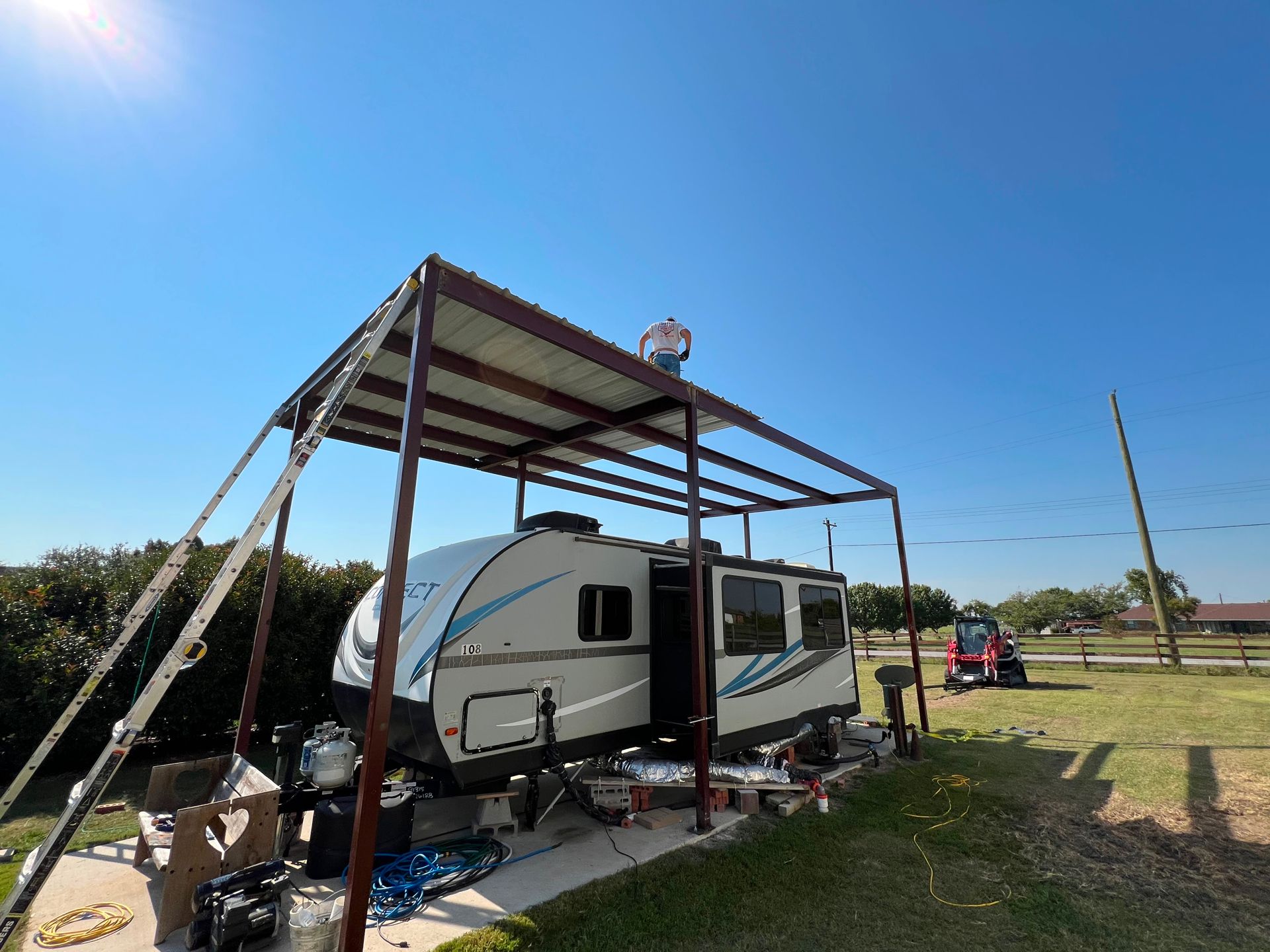 A person on a metal carport roof above a white and blue travel trailer on a sunny, grassy lot.
