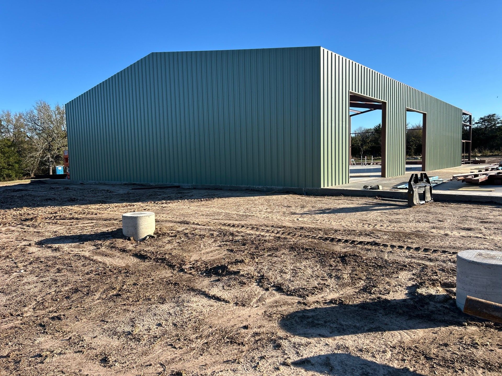 A green corrugated metal building under construction on a dirt lot with two concrete pillars in the foreground.