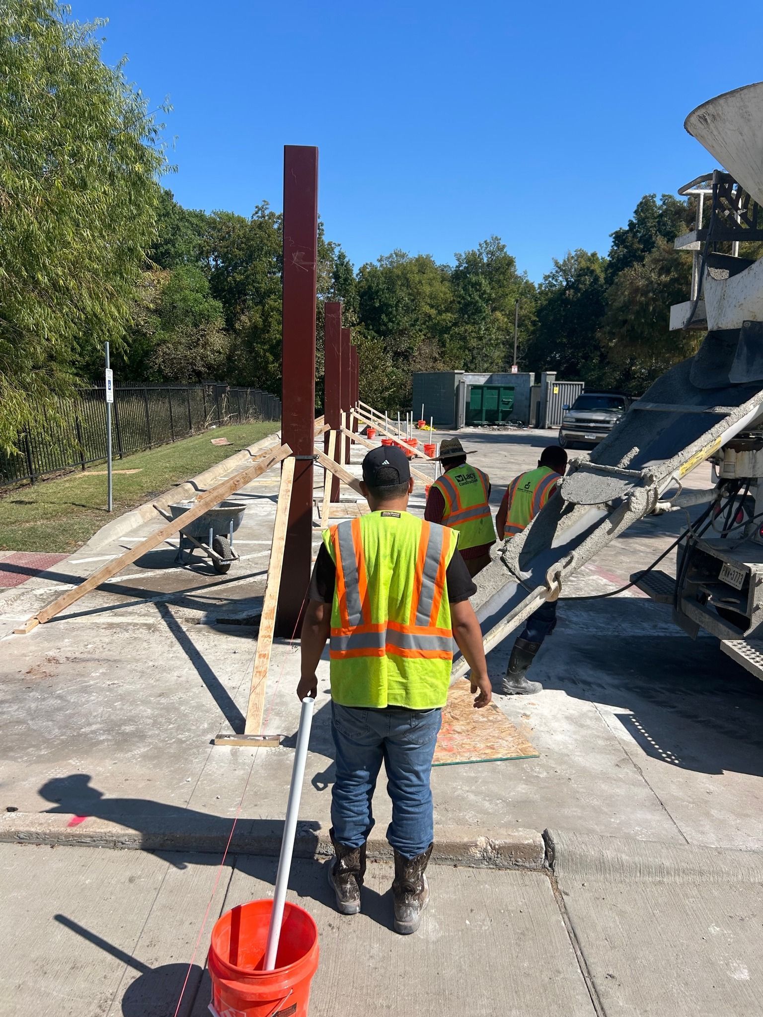 Construction workers pour concrete around vertical steel posts at an outdoor job site.