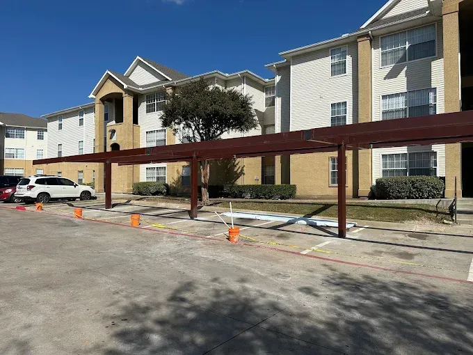 A partially constructed brown metal car port structure in the parking lot of an apartment complex with orange safety cones.