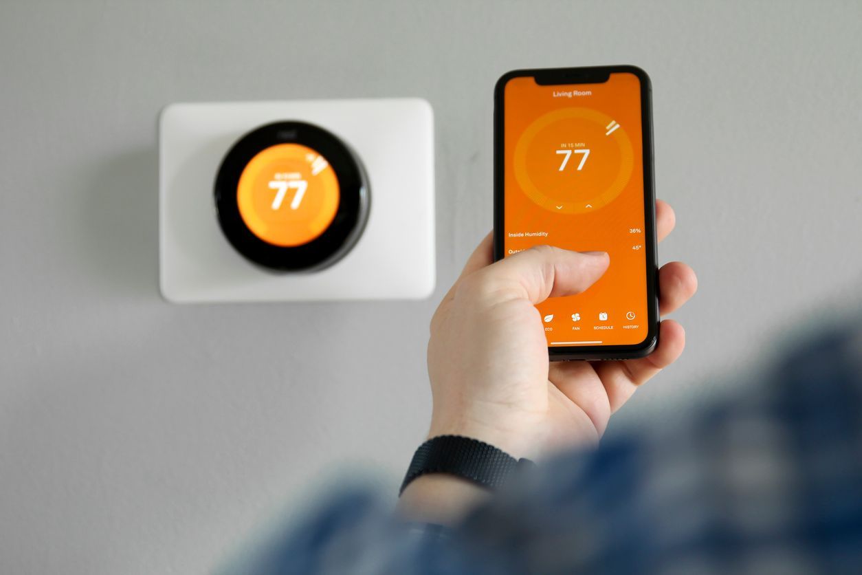 Hand holding smartphone controlling a wall thermostat, both showing 77° on orange screens