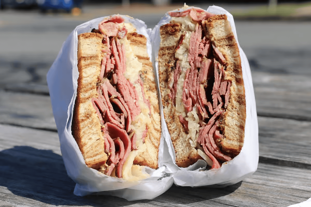 Large pastrami sandwich, cut in half, wrapped in white paper, on a wooden table.