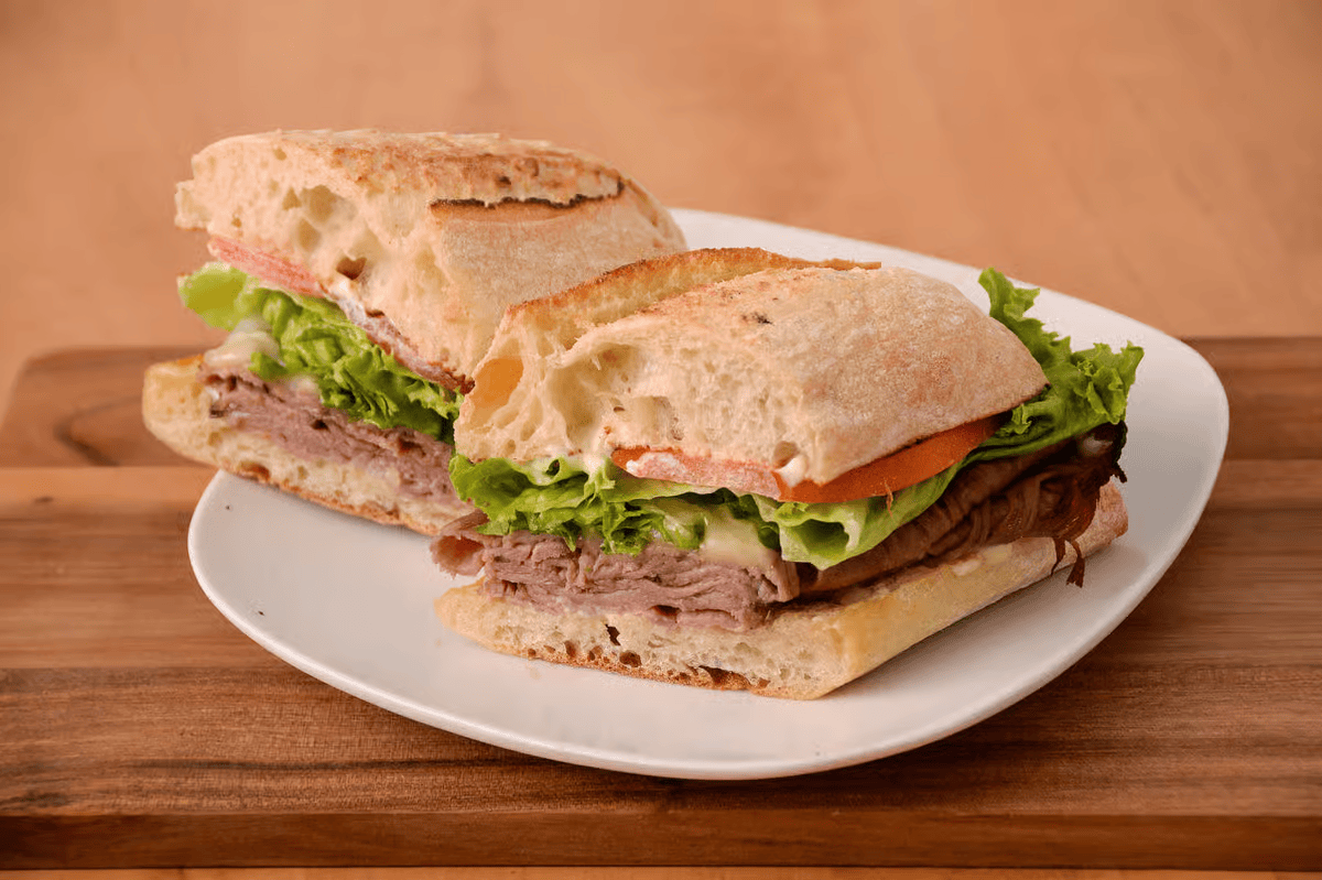 Sliced sandwich on a white plate, showcasing layers of meat, lettuce, and tomato on crusty bread, resting on a wooden surface.
