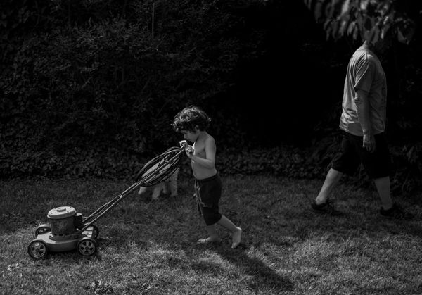 A child pushes a small lawn mower across a grassy yard while an adult walks nearby in a black-and-white scene.