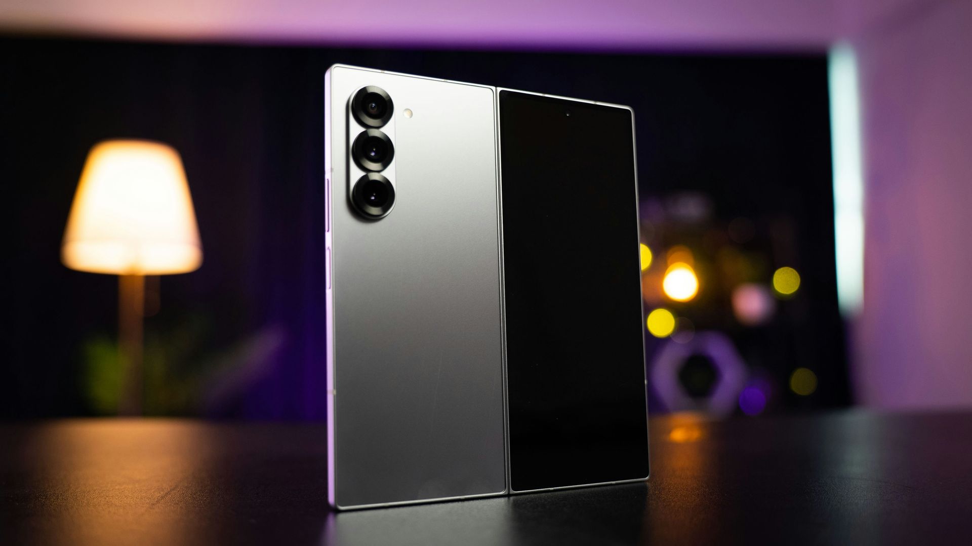 Silver foldable smartphone standing on a table with a blurred lamp and lights in the background