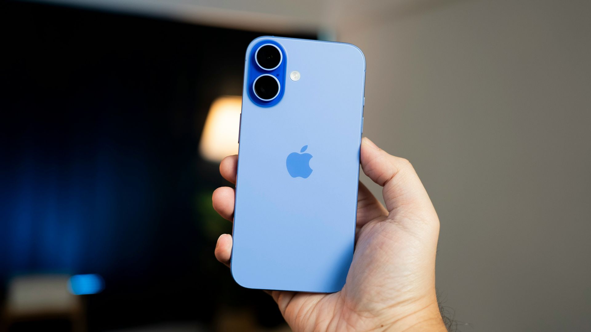 Hand holding a blue iPhone with dual cameras against a blurred indoor background