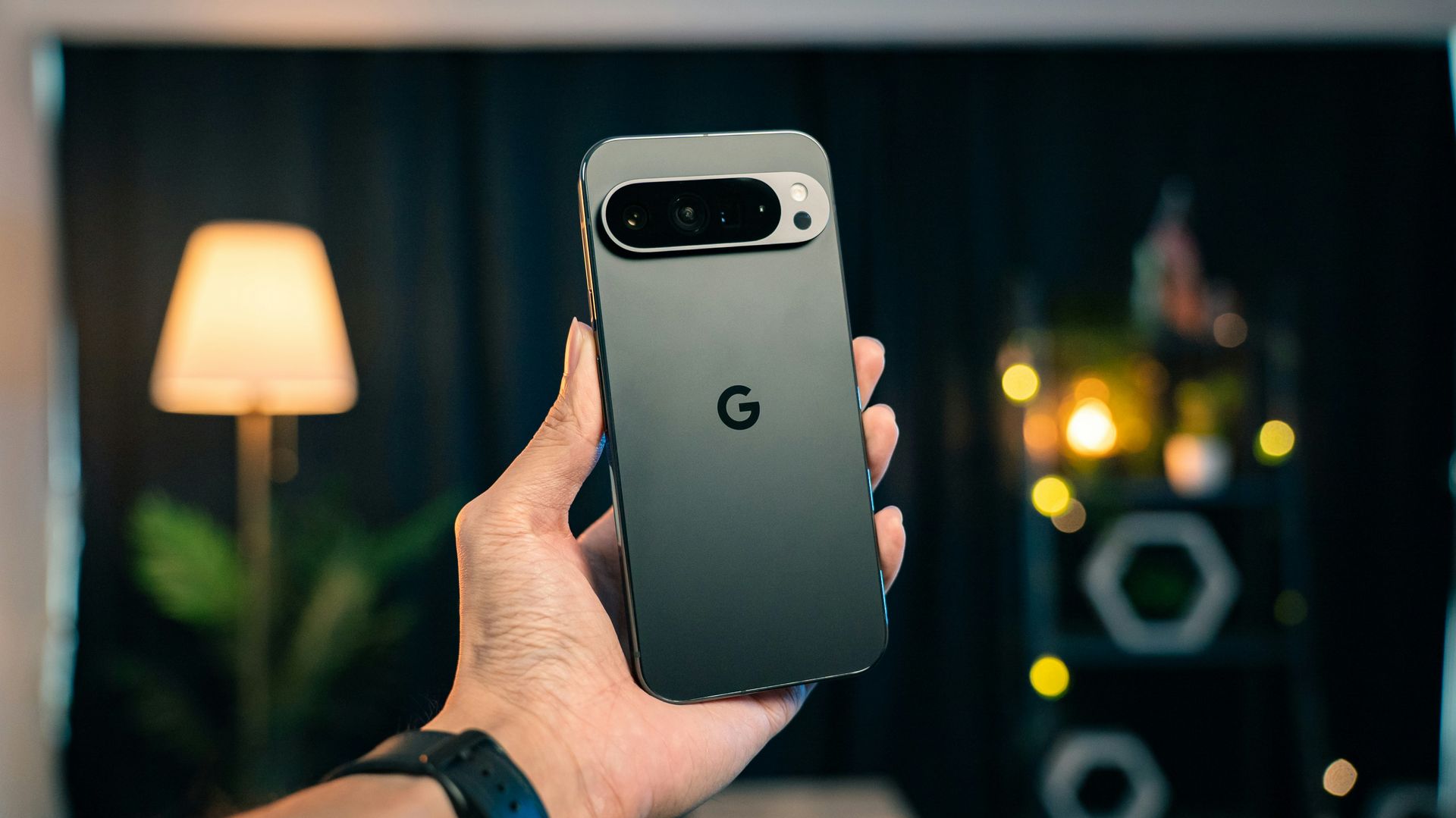 Hand holding a dark Google Pixel phone with a Google logo, indoors with a lamp and blurred lights in background