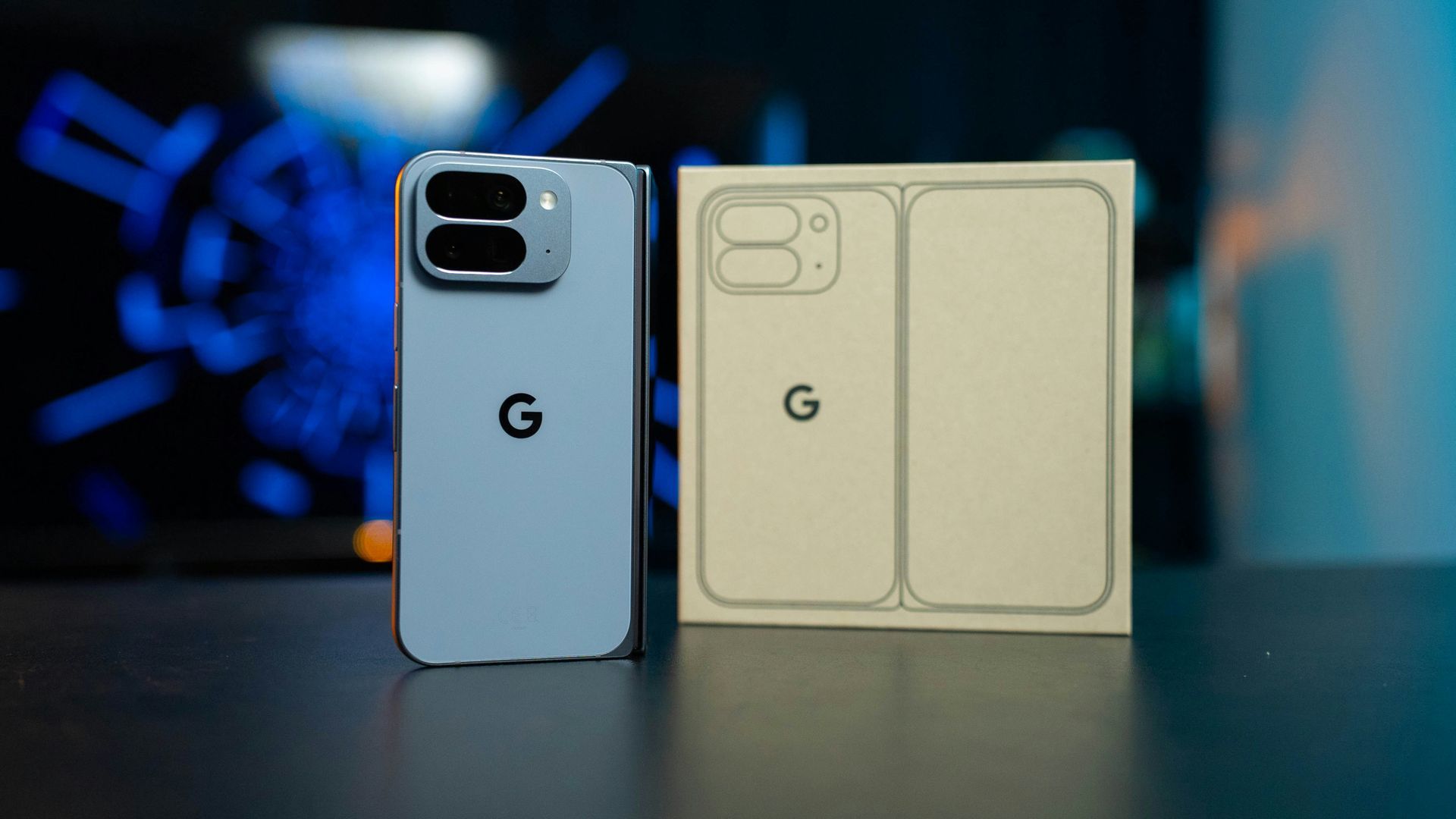 Two Google Pixel phones standing side by side, one blue and one beige, on a table with blurred blue lights behind.
