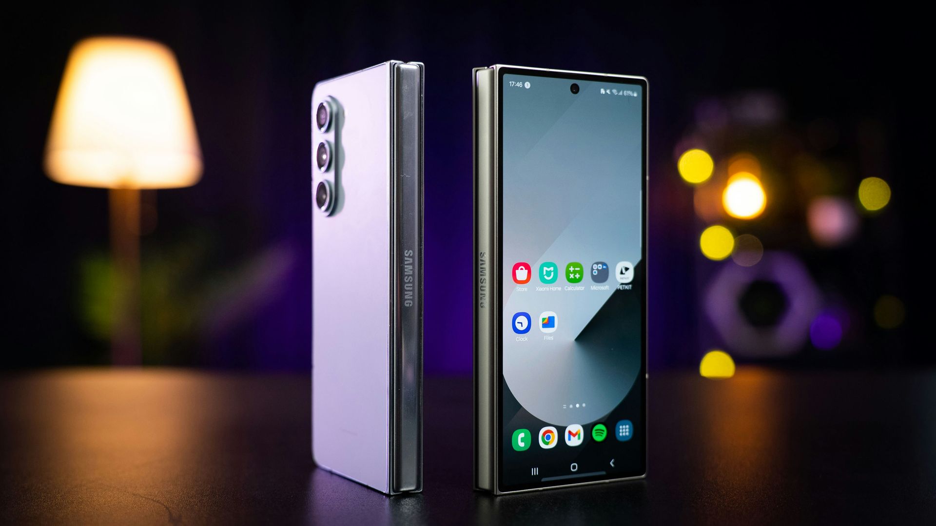 Foldable smartphone standing open, showing front screen and silver back on a dark table with blurred lights behind