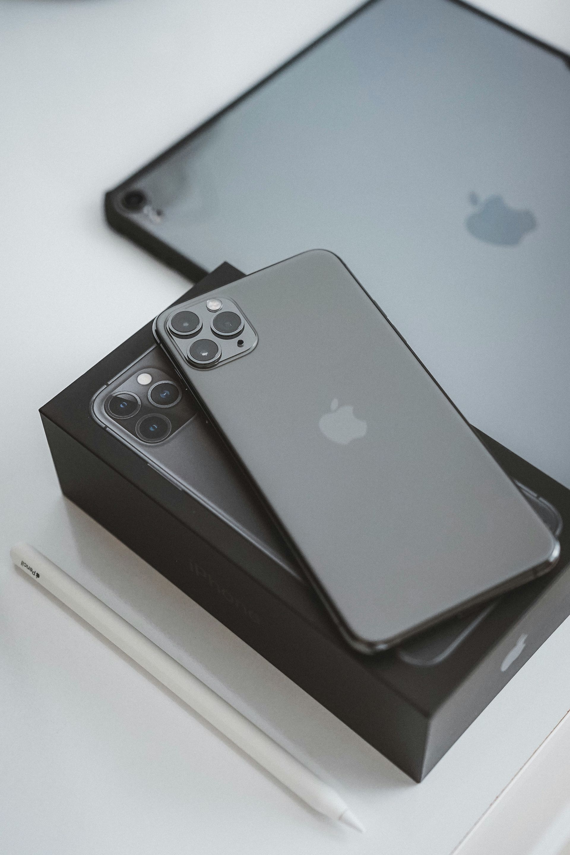 Three gray Apple devices stacked on a black box, including two iPhones and an iPad.
