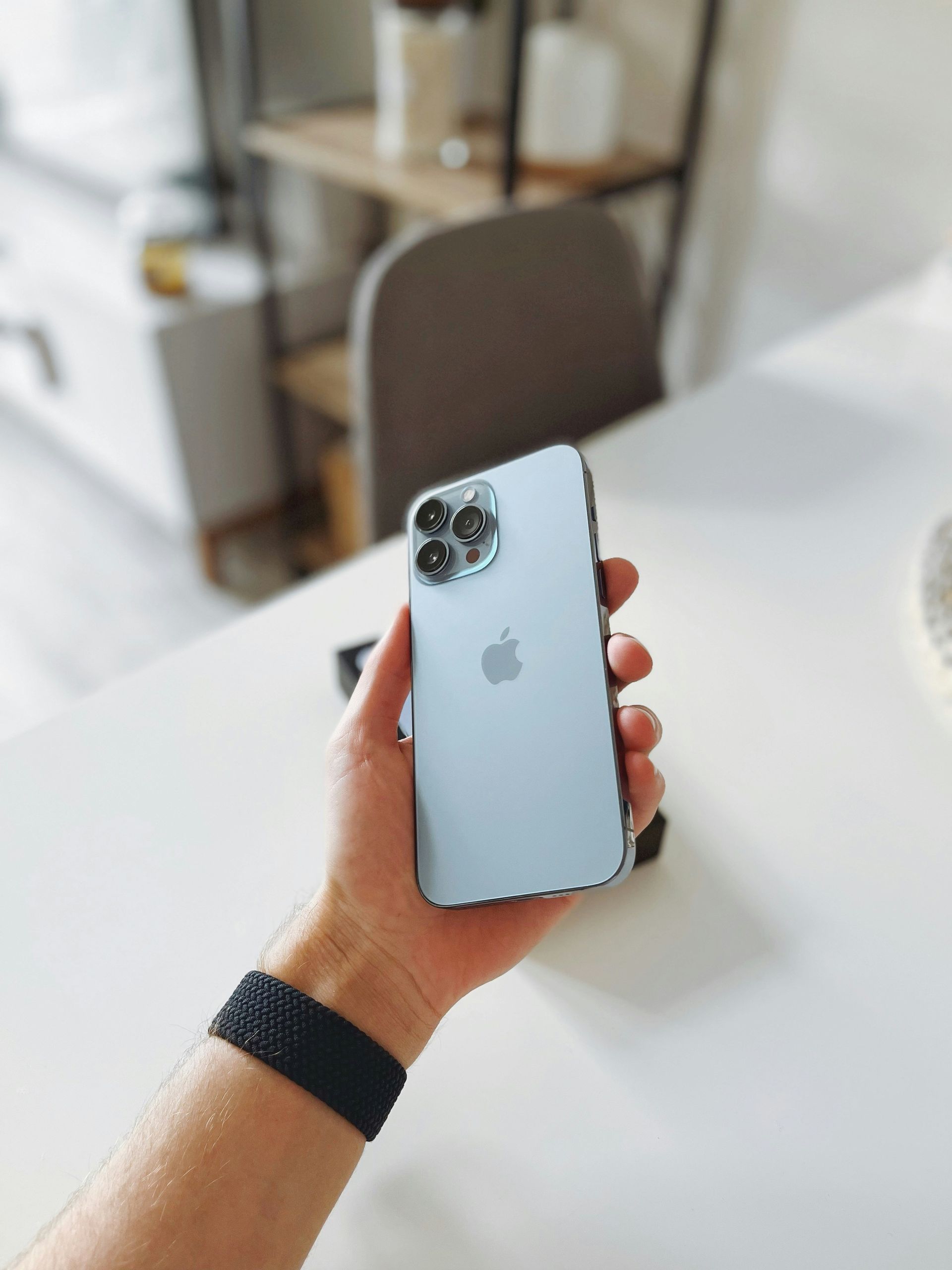 Hand holding a silver iPhone with triple cameras over a white table in a bright room