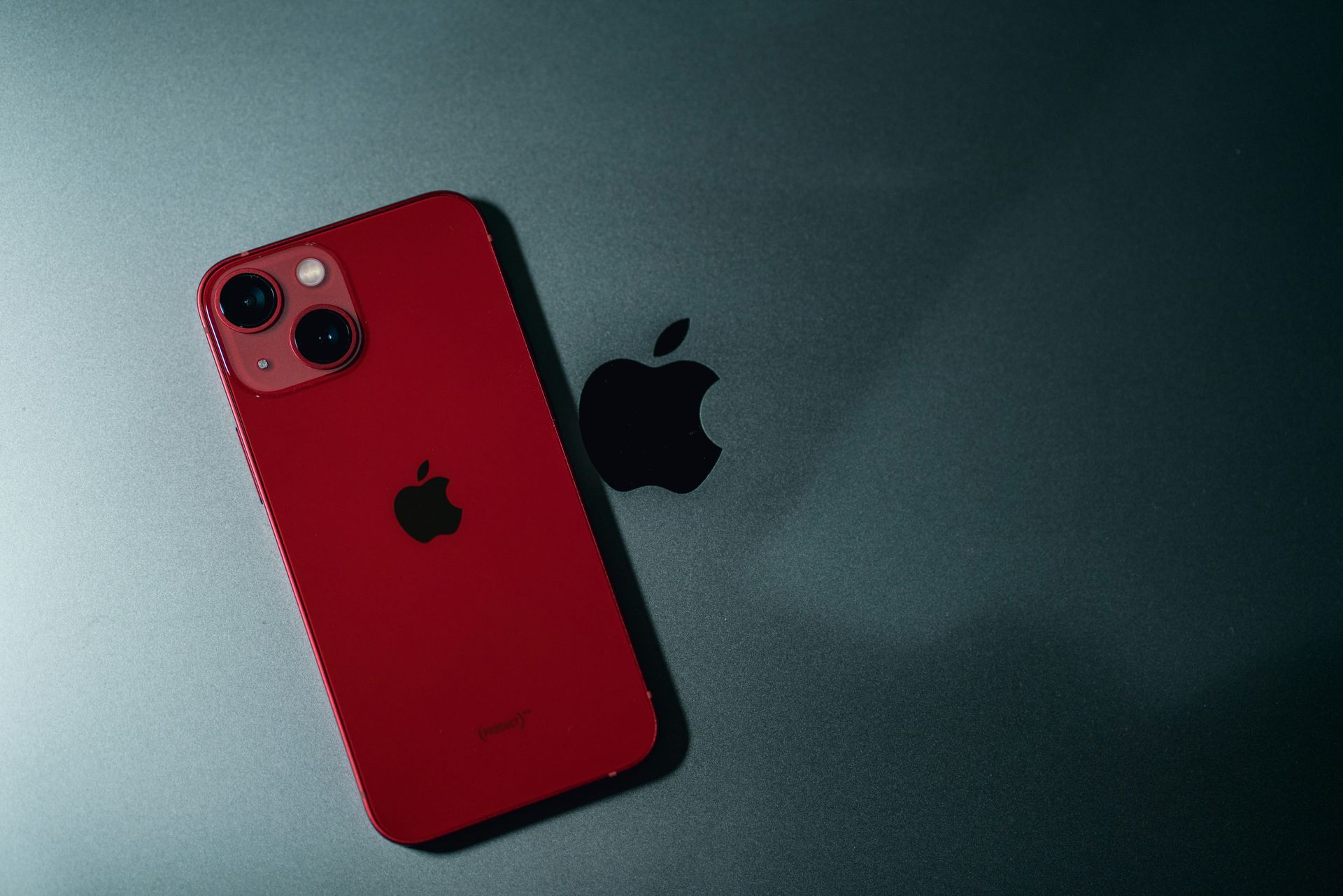 Red iPhone on a dark surface beside a black Apple logo.