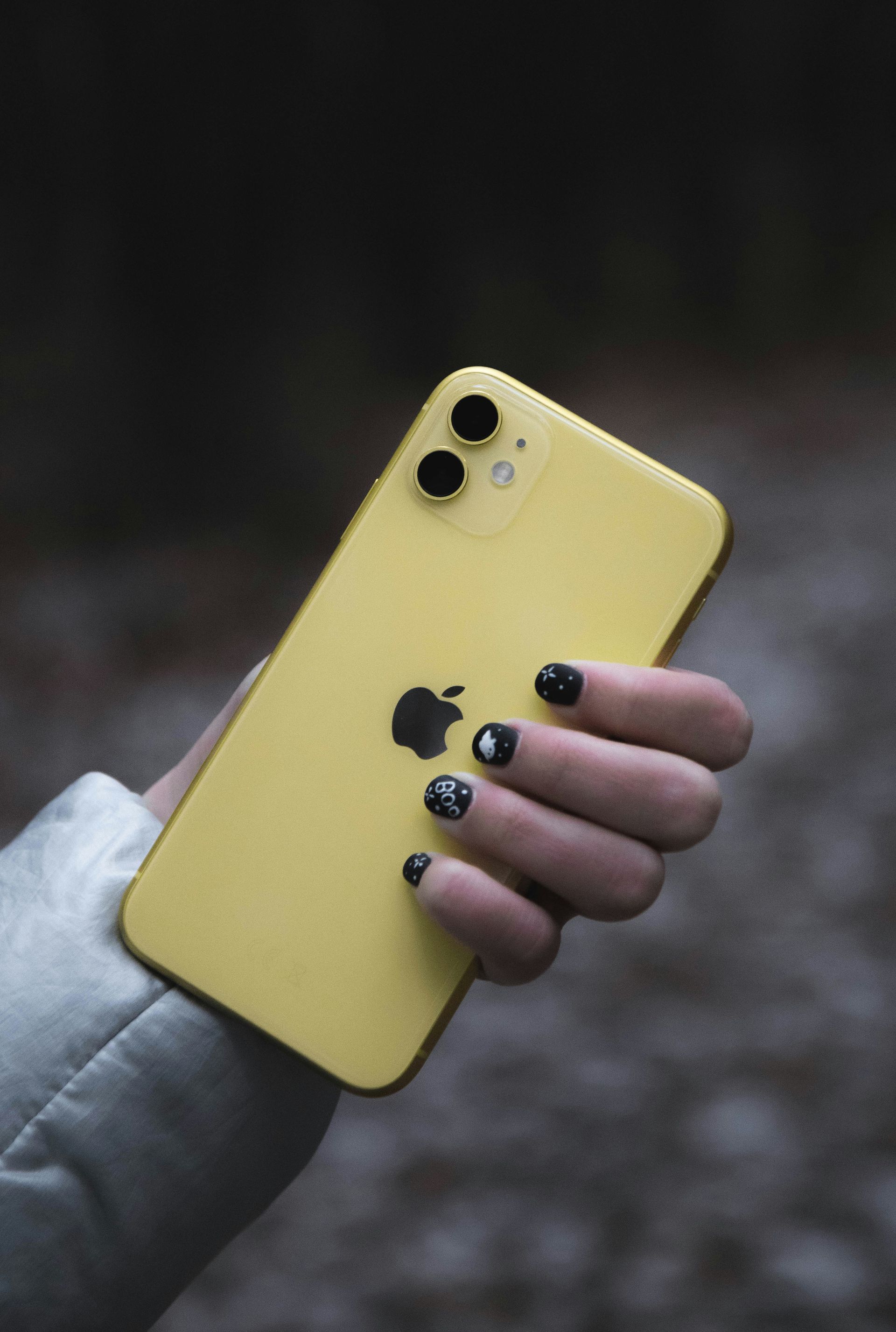 Hand holding a yellow iPhone with black nails against a blurred outdoor background