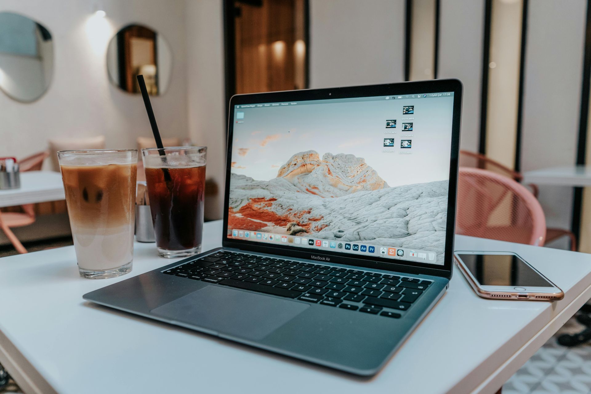 Laptop on a café table beside iced drinks and a phone, with a desert wallpaper on screen.