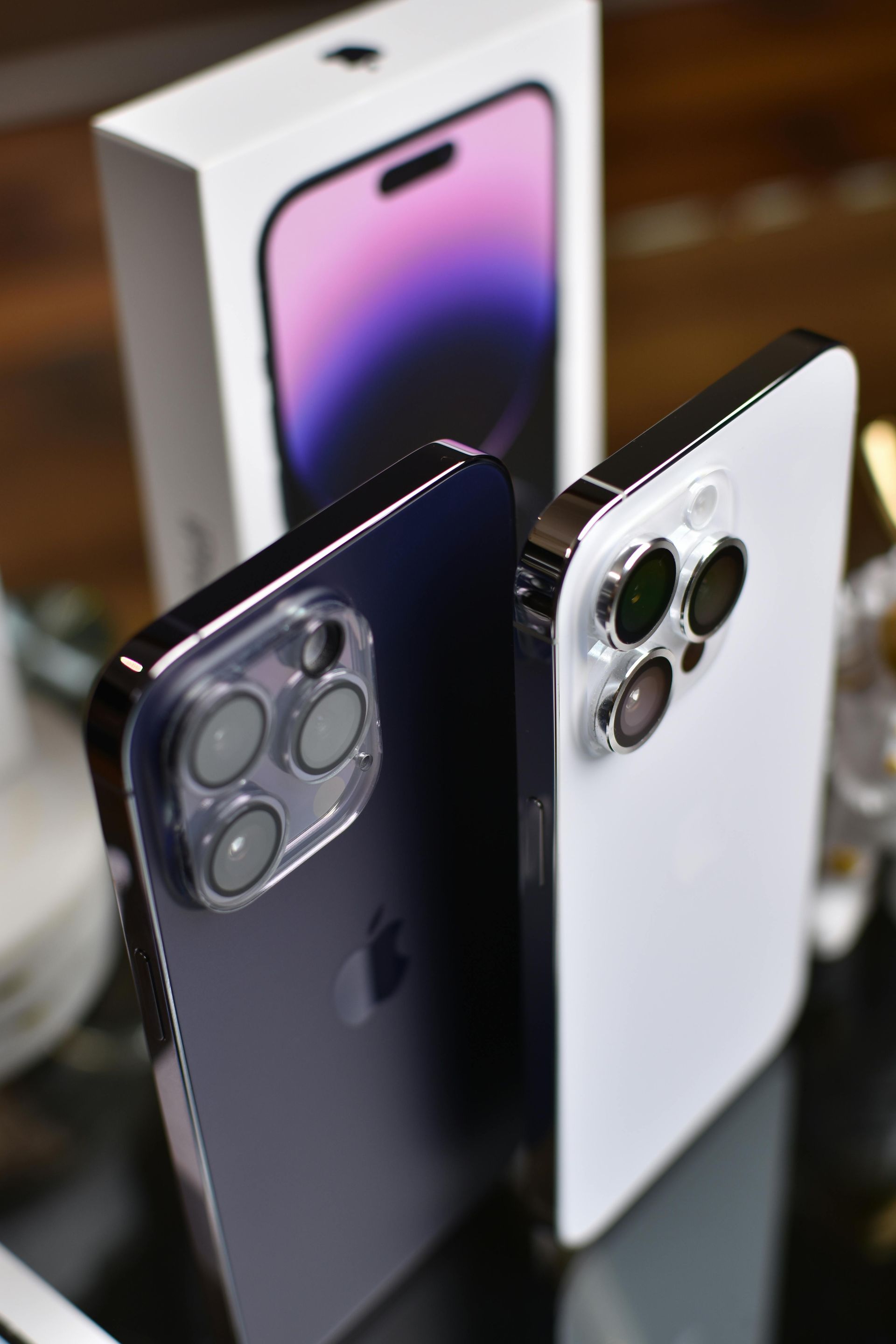 Two iPhones in purple and white rest on a reflective surface in front of their product box displaying the purple model.