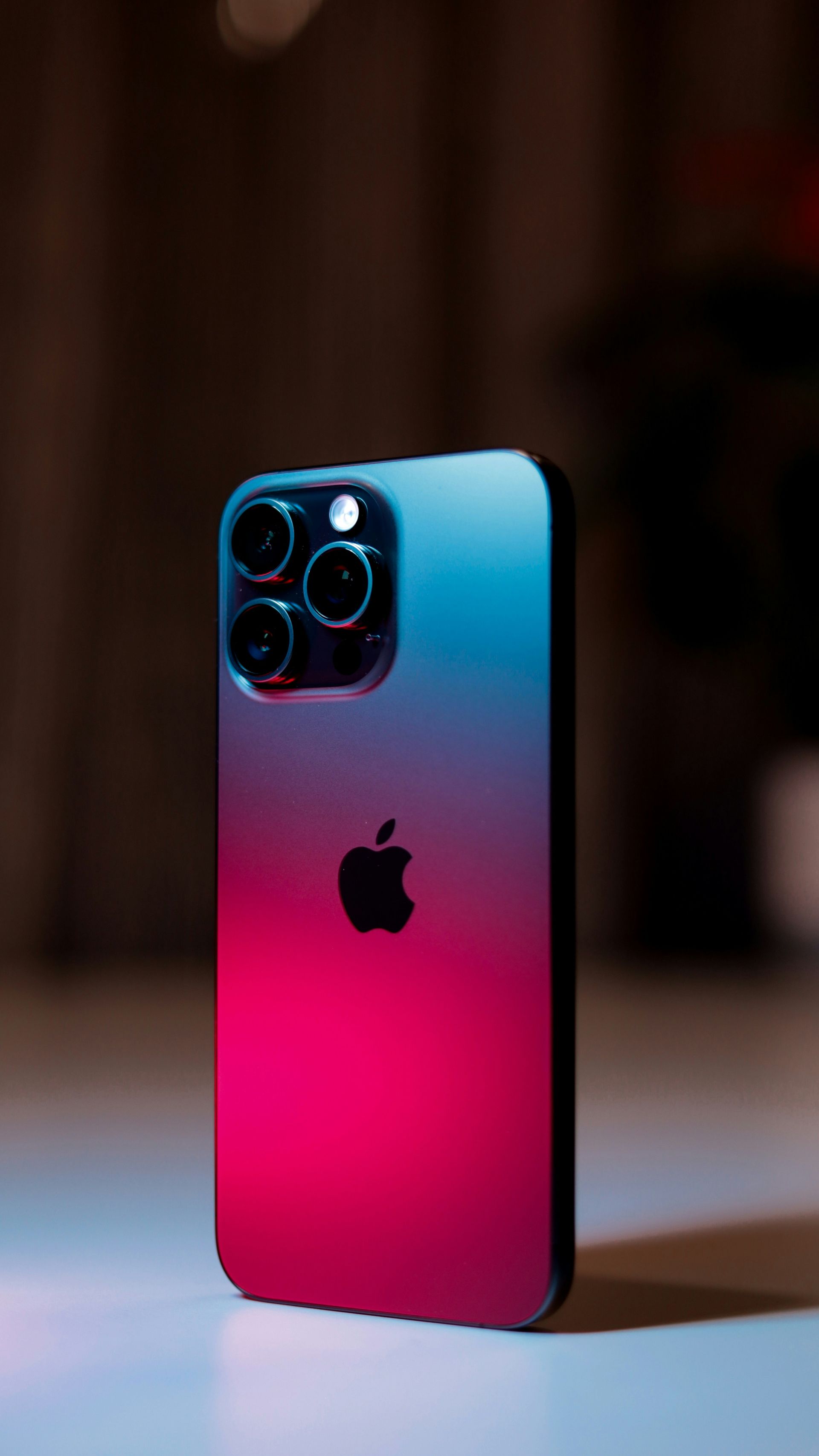 Blue-to-pink gradient iPhone with triple camera module standing upright on a table