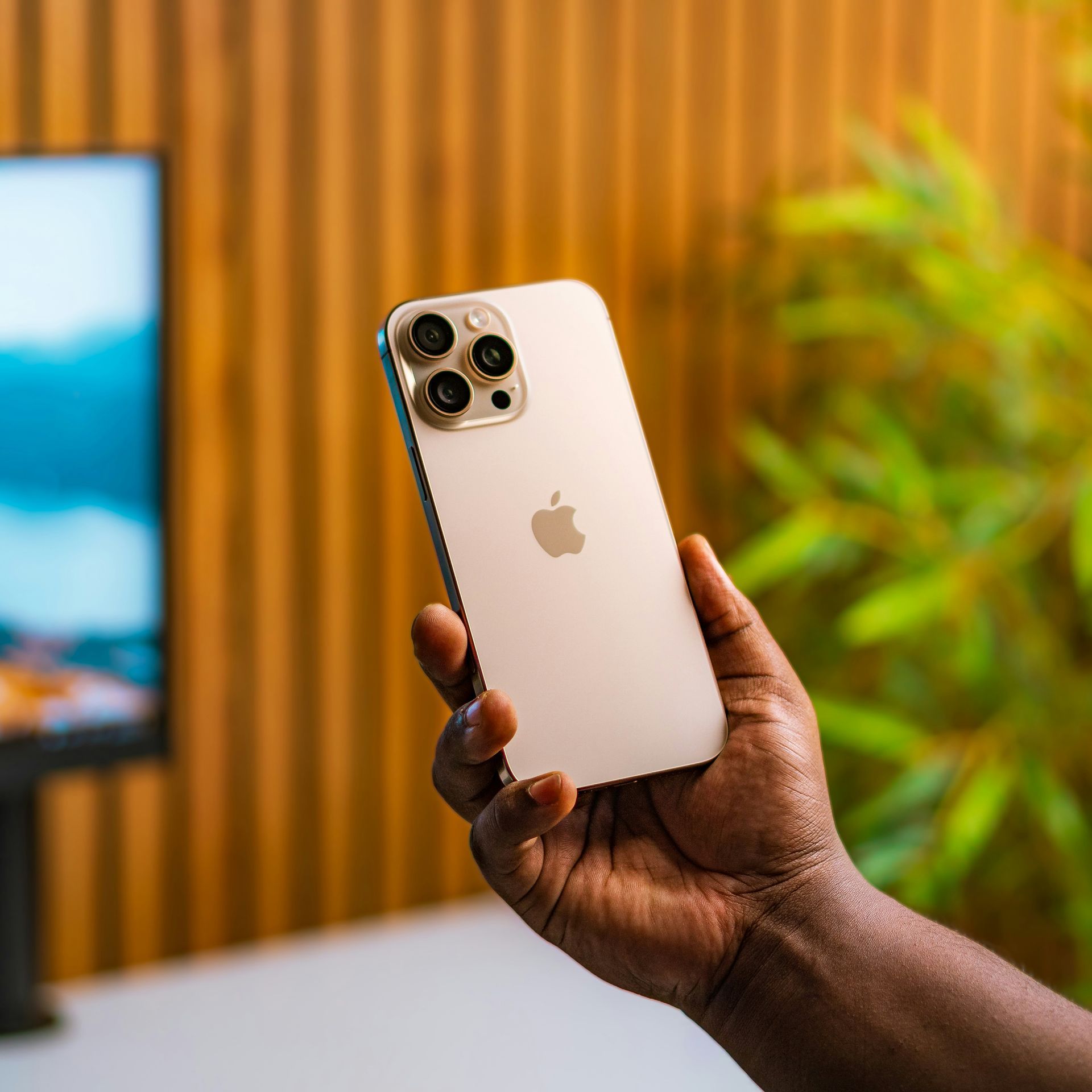 Hand holding a gold iPhone with triple cameras, against a blurred wood-paneled background
