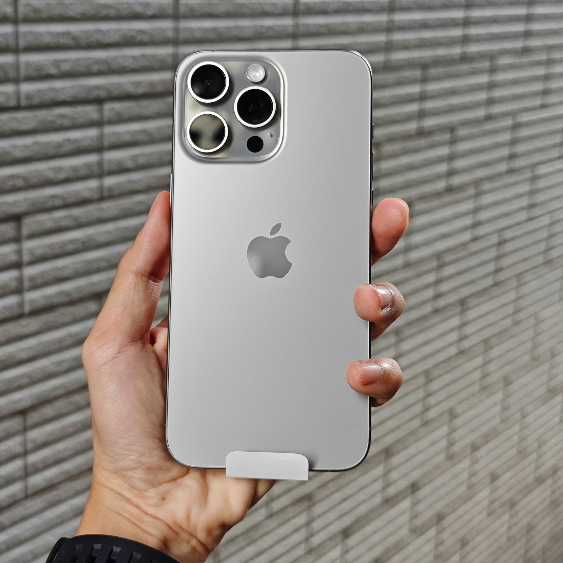 Hand holding a silver iPhone with three rear cameras against a gray tiled wall.