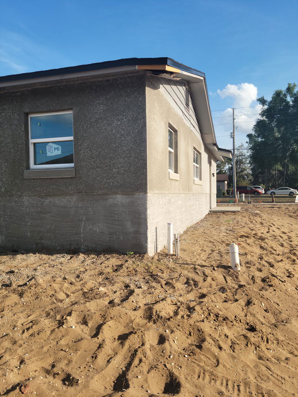 Corner of a new, gray stucco house under construction with a sandy yard and clear blue sky.