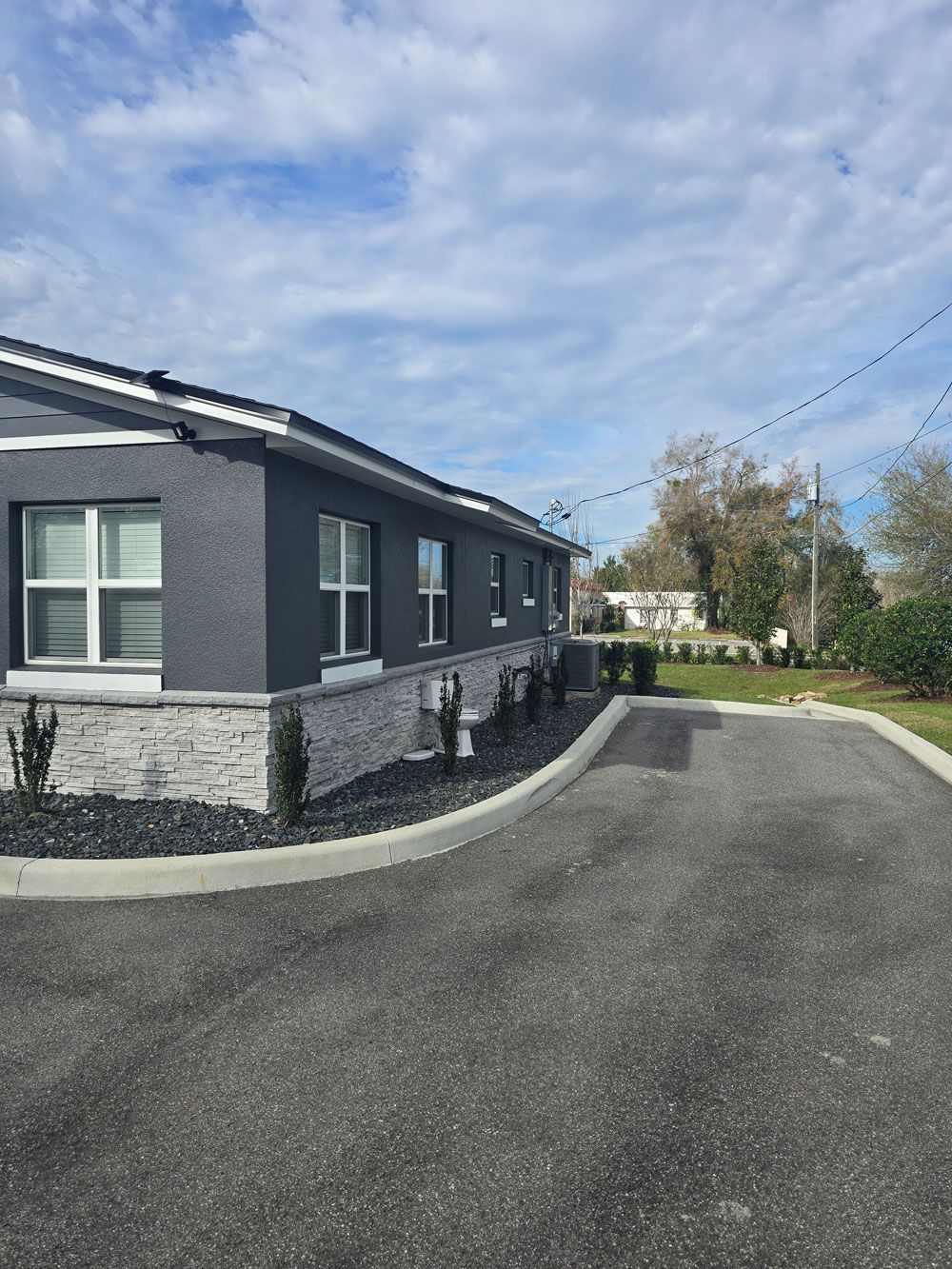 Exterior view of a dark gray building with a stone veneer base, black asphalt driveway, and a blue sky overhead.