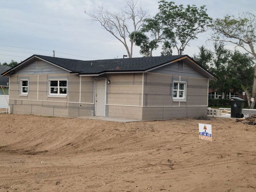 A partially constructed house with a dark roof, light brown siding, and white window frames, in a dirt lot.